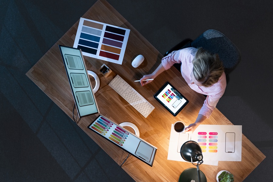 Overhead view of an individual at a desk with 2 monitors and a tablet. Documents are also random locations on the desk.
